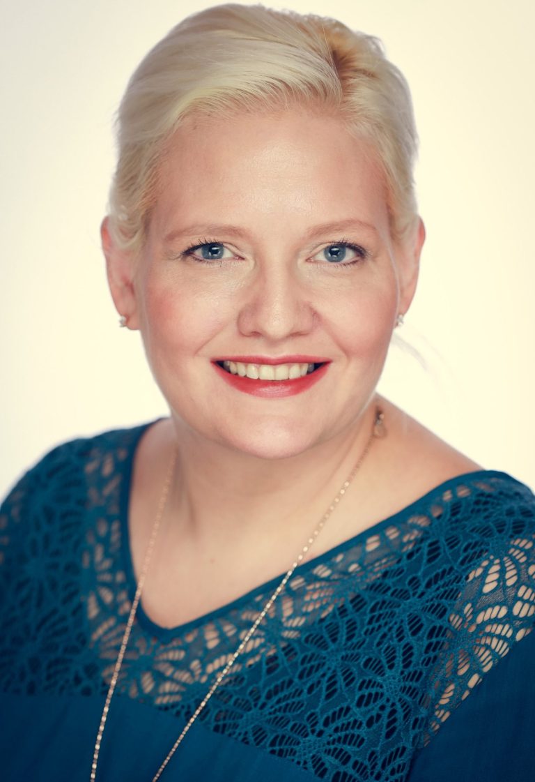 Eine Frau mit blonden Haaren, die sie zusammen gebunden trägt, lächelt freundlich in die Kamera.