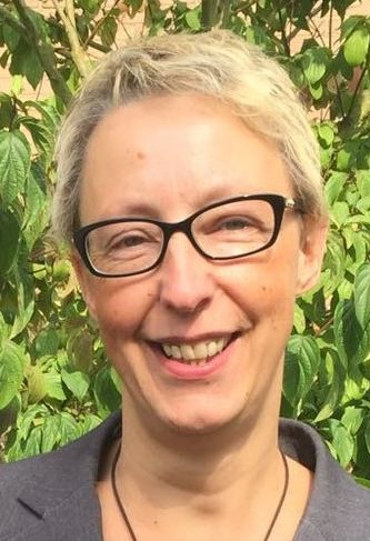 Eine Frau mit blonden, kurzen Haaren lächelt freundlich in die Kamera. Sie steht vor einer grünen Pflanze.